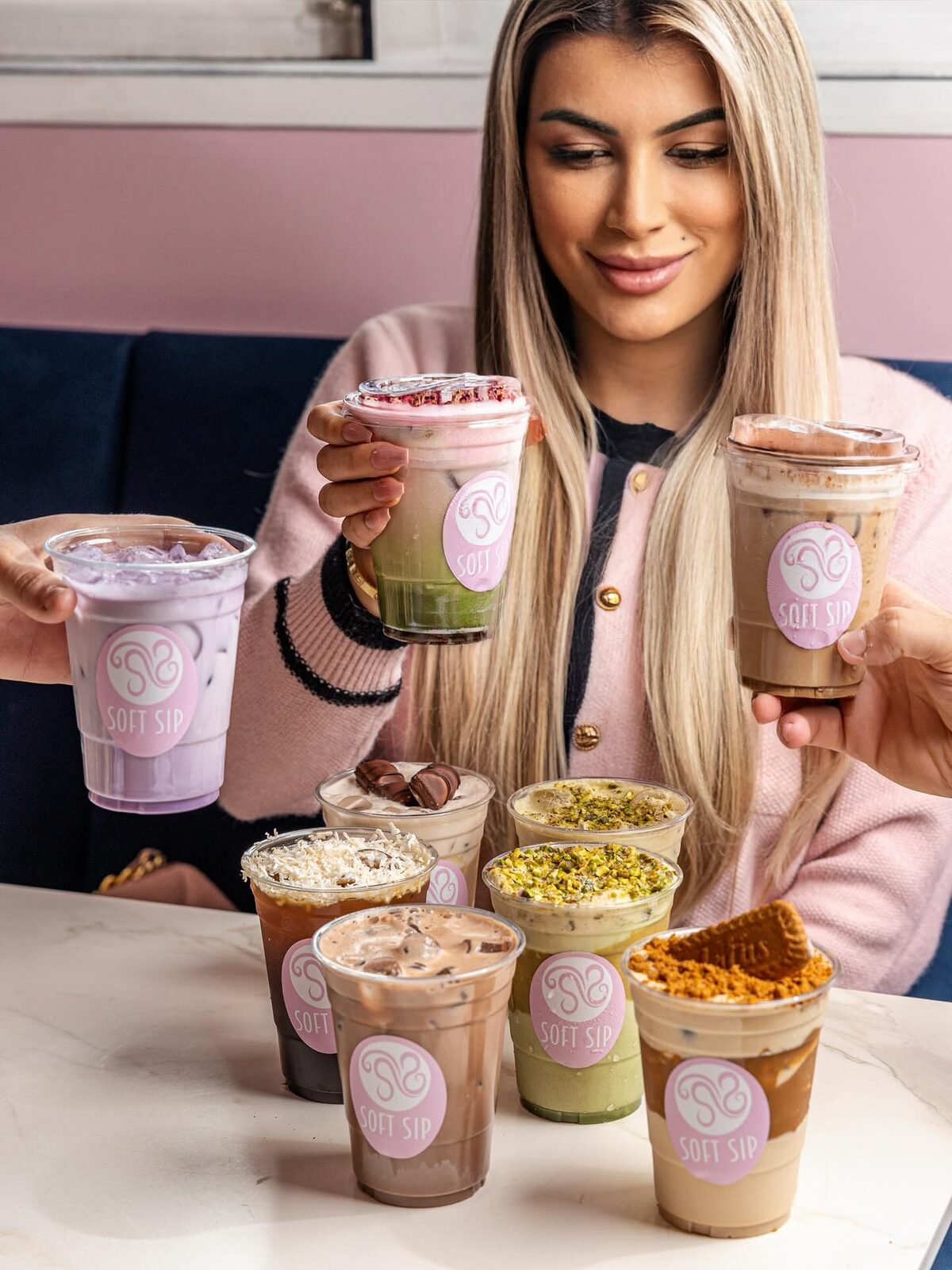A customer holding three Soft Sip signature iced drinks with more lined up on the table: pink, matcha, caramel and chocolate layers